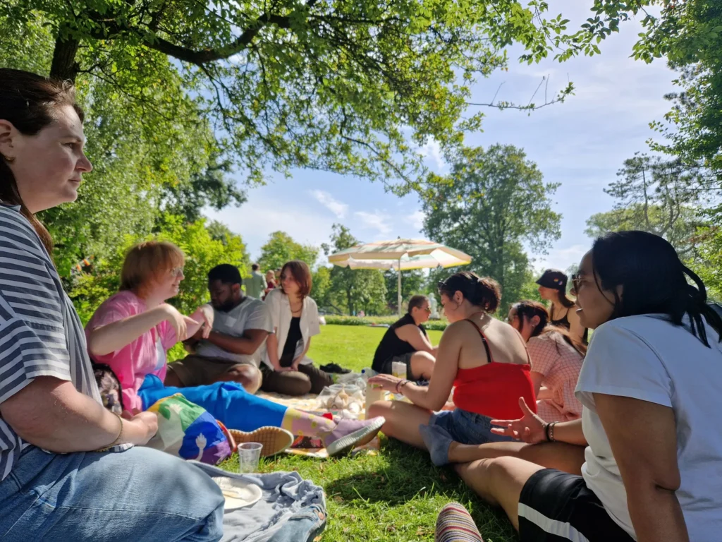 A gathering of people at a park, people in the back are having a picnic.