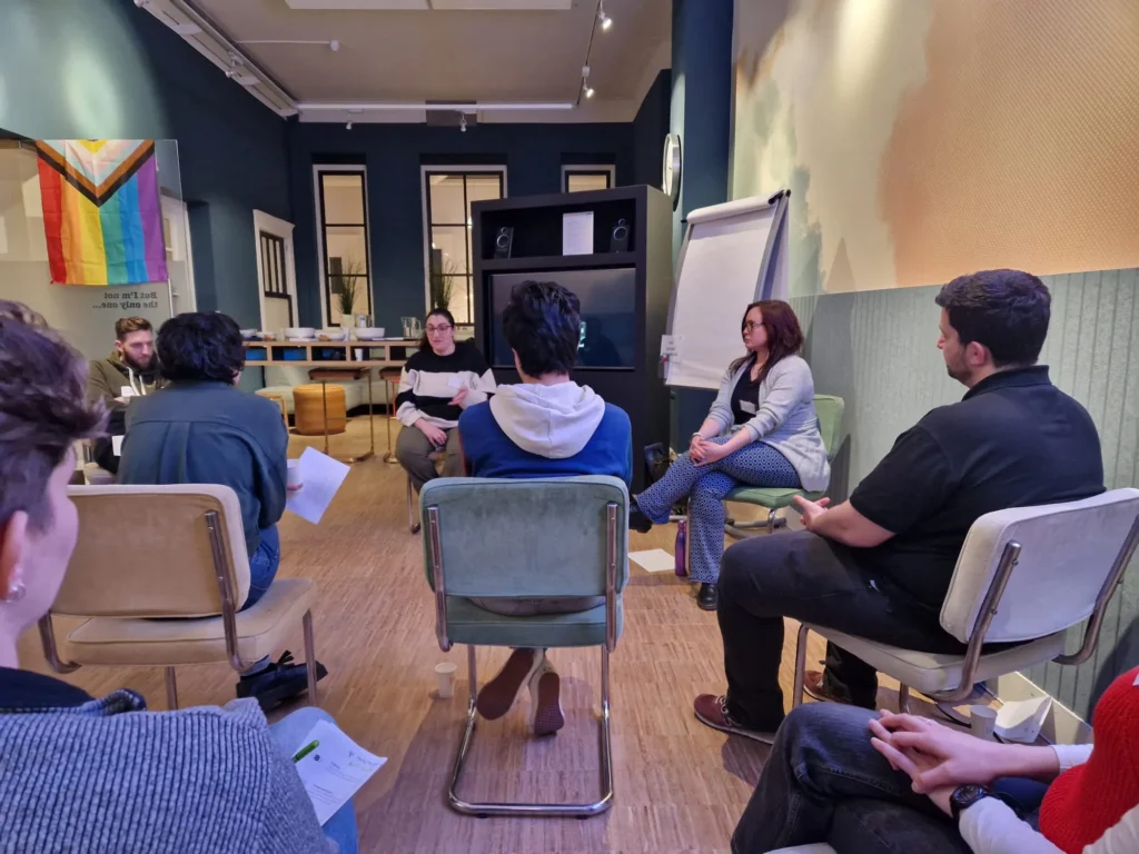 People sitting in a circle, having a meeting. In the background is a pride flag and a flipover.