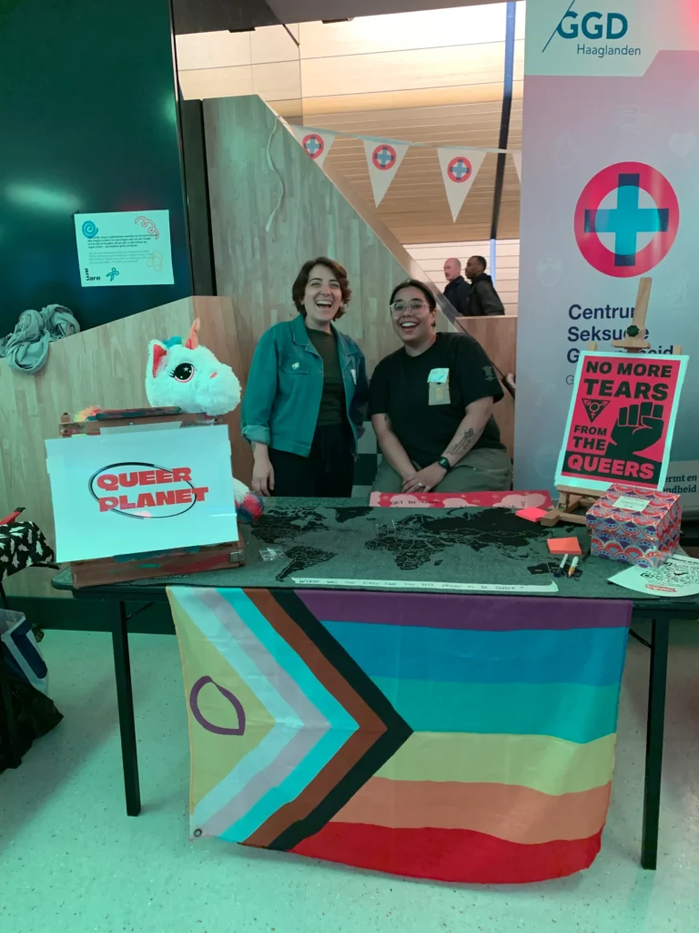 Two people standing behind a table dressed in Queer Planet logos, a pride flag and a map of the world. One of the signs says "No More Tears for the Queers".